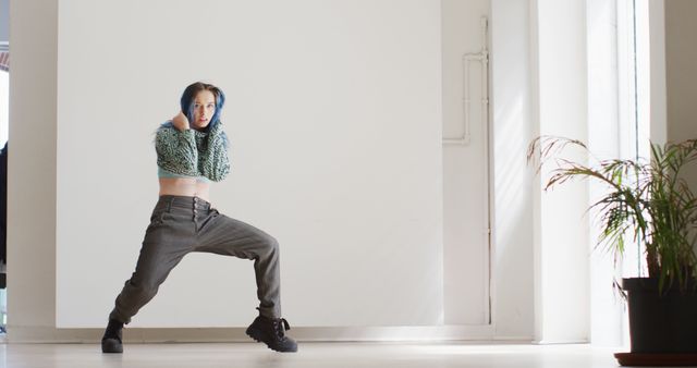 Young woman with blue hair energetically dancing indoors in funky casual clothes. Useful for promoting urban fashion, dance classes, and expressing modern lifestyles.