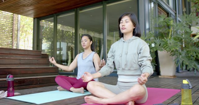 Two women sitting cross-legged and meditating on yoga mats on a wooden deck, providing a serene outdoor environment ideal for relaxation and mindfulness. Suitable for use in content related to health and wellness, yoga practices, mental health awareness, or lifestyle blogs promoting self-care routines.