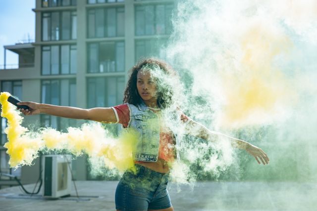 Front view of a hip young biracial woman wearing denim jacket and jeans, holding a smoke grenade with yellow smoke on an urban rooftop with building in the background