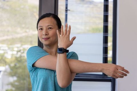 Asian woman stretching arms indoors wearing smartwatch for fitness and wellness routine
