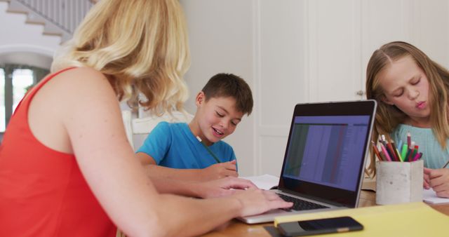 Mother homeschooling two children at dining table with laptop and educational supplies. Kids are engaged in assignments while mother guides them. Ideal for educational content, homeschooling resources, family education, and distance learning promotion.