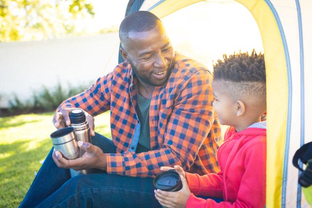 Father and son sitting in a backyard tent, sharing hot drinks from a thermos. Perfect for themes related to family bonding, outdoor activities, parenting, and leisure time. Ideal for use in advertisements, blogs, and articles about family life, camping, and spending quality time together.