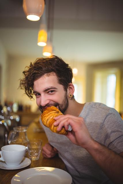 Young man enjoying a croissant at a trendy cafe. Ideal for use in lifestyle blogs, breakfast menus, and social media content related to food, cafes, and relaxed lifestyle.