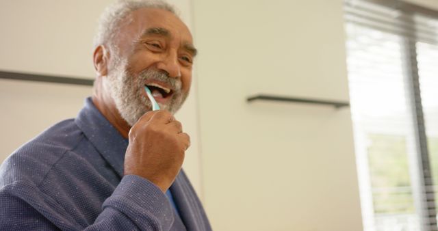 This image shows a smiling African American senior man brushing his teeth in a well-lit bathroom. Ideal for use in healthcare promotions, dental care advertisements, wellness articles, and marketing materials targeting senior health and healthy living habits.
