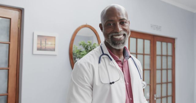 Smiling African American male doctor wearing a white coat and stethoscope in a clinical office setting. Ideal for promoting healthcare services, medical websites, patient care pamphlets, and advertisements for hospital or clinic services.