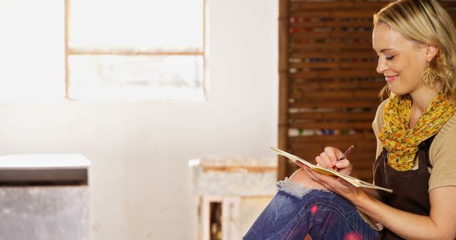 Smiling female artist painting with passion in bright, sunlit studio. Natural light illuminates her workspace, creating a warm and motivating environment. Ideal for promoting art workshops, highlighting creative processes, or picturing a tranquil and creative environment.