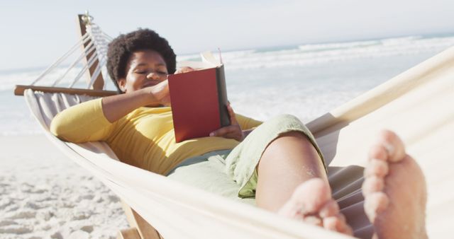 A woman is lying in a hammock on a sunny beach, enjoying a book. The waves are gently crashing in the background. This image is perfect for promoting travel destinations, leisure activities, relaxation techniques, or book clubs.