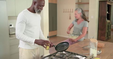 Mature couple preparing breakfast pouring oil into frying pan on kitchen island