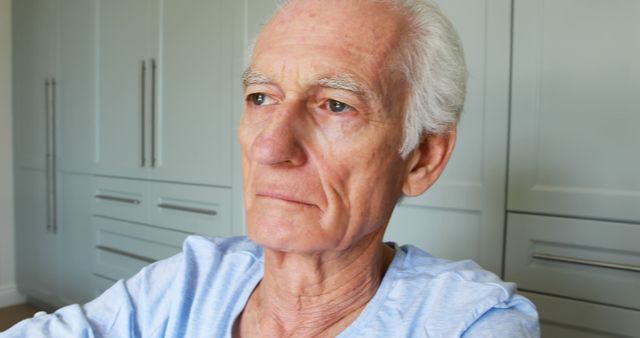 Elderly man with white hair wearing a light blue shirt, sitting and looking thoughtful. Background includes light-colored cabinets, indicating a cozy home environment. Perfect for use in articles or advertisements related to aging, retirement, health care, senior living, and personal contemplation.