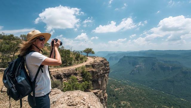 Female hiker photographing dramatic canyon from cliff rim with backpack and sun hat