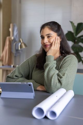 Young woman contemplating design ideas at home studio with tablet and rolled blueprints