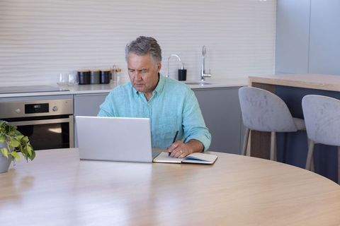 Senior Man Working from Home at Kitchen Table Using Laptop, Writing in Notebook