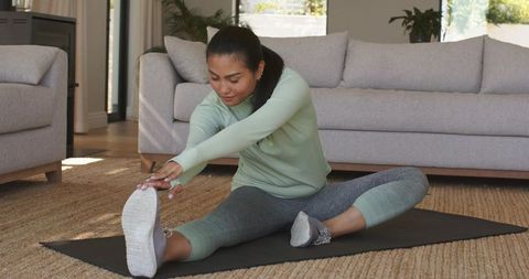 Asian woman stretching on mat in living room wearing green leggings for home wellness