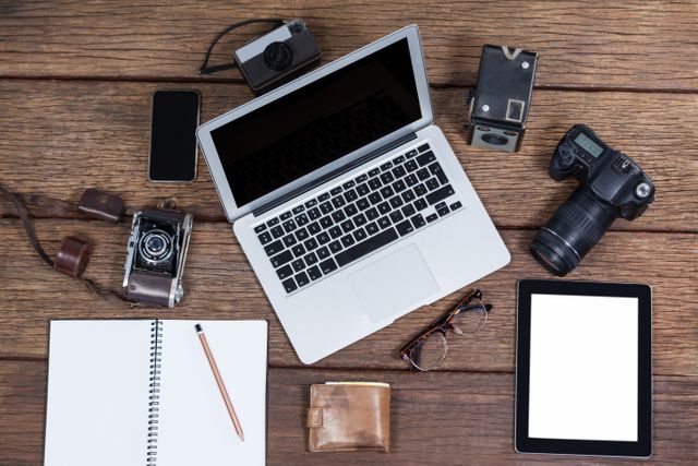 This image depicts a creative workspace featuring a laptop, various vintage cameras, a smartphone, a tablet, a notebook with a pencil, glasses, and a wallet on a wooden table. Ideal for use in articles or advertisements related to photography, creative workspaces, technology, or vintage equipment. Perfect for blogs, websites, or social media posts highlighting the blend of modern and vintage technology.