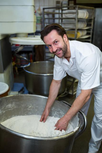 Baker is mixing flour in an industrial kitchen, wearing a white uniform and smiling at the camera. Ideal for use in articles or advertisements related to baking, culinary arts, professional kitchens, or the food industry. Can be used to depict a positive and professional work environment in a bakery or culinary setting.