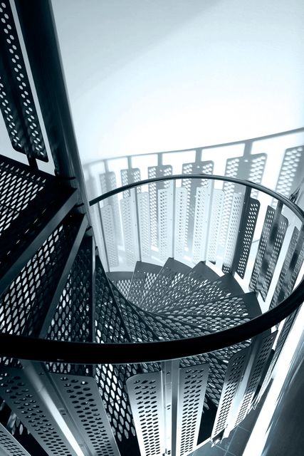 Photograph shows a modern spiral staircase with a geometric pattern, focusing on the abstract design and play of light and shadows. Ideal for use in architectural design websites, modern interior design portfolios, or abstract artwork collections.