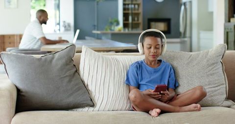 African American Father Working Laptop While Son Wearing Headphones and Using Phone in Living Room