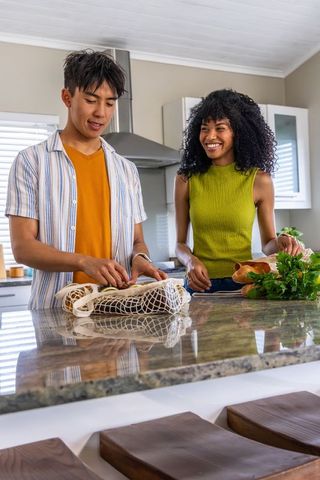 Black woman and asian man unpacking groceries at modern kitchen island, bright daylight