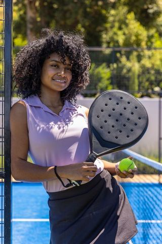 Smiling african american woman leaning on fence holding padel racket and ball on sunny court