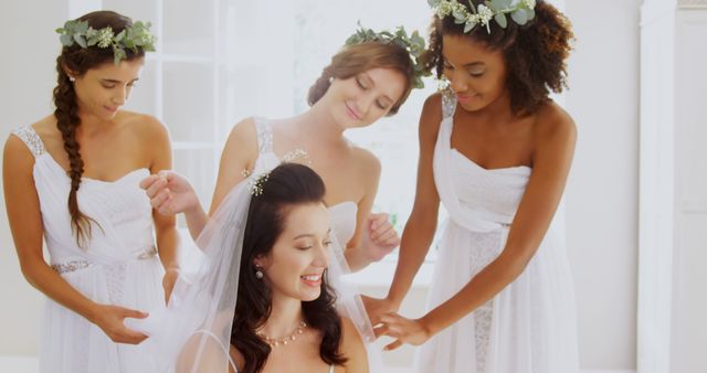 Bride having dress and veil arranged by bridesmaids who are smiling and wearing white dresses and flower crowns in a brightly lit room. Useful for wedding planning, bridal preparation, beauty and fashion editorials, and friendship themes.