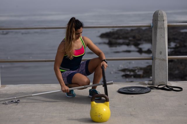 Caucasian woman with long dark hair wearing sportswear preparing barbells for strength training by the seaside on a sunny day. Ideal for use in fitness blogs, workout tutorials, outdoor exercise promotions, and health and wellness articles.