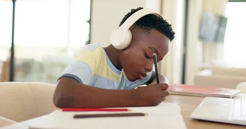 African american boy studying at home wearing headphones writing homework at desk focused