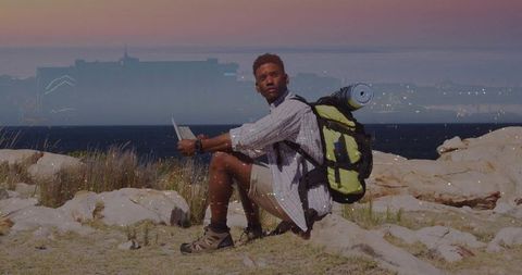 Sitting hiker checking tablet on coastal rocks at dusk with backpack and rolled mat