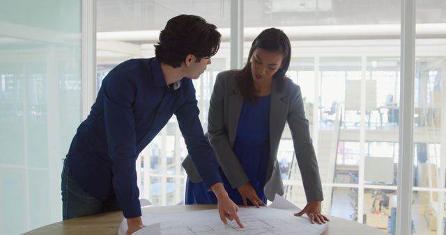 Businessman and businesswoman review plans in a bright office. They collaborate over architectural blueprints, indicating teamwork and strategy.