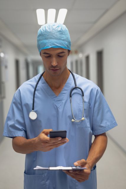 Male surgeon in blue scrubs and cap using mobile phone in hospital corridor. Stethoscope around neck, holding clipboard. Ideal for healthcare, medical technology, communication, and modern medicine themes.