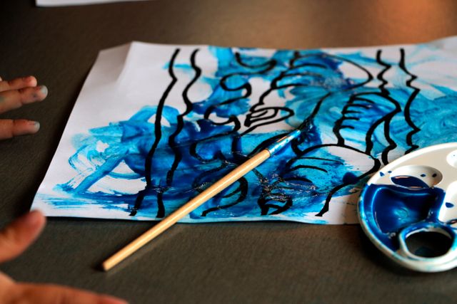 Abstract blue ink painting on paper with child's hands partially visible. Features a wooden brush and a palette with blue ink. Ideal for illustrating children's creativity, art classes, kid's artistic expression, or home art projects.