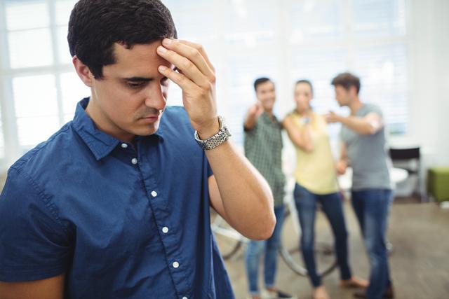 Man in blue shirt appears stressed while colleagues in background are teasing him. Useful for illustrating workplace bullying, emotional distress, and the importance of mental health in professional settings.