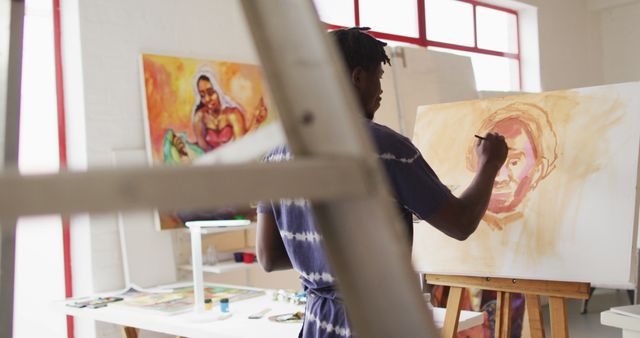 Male artist painting a portrait on a canvas in a bright art studio filled with colorful paintings. Perfect for use in articles or websites related to art, creativity, artist biographies, workshops, and creative spaces.