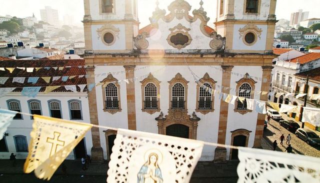 Sunlit baroque church facade with papel picado banners casting warm shadows over plaza