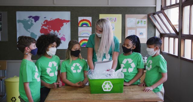 Teacher and young students in green recycling shirts gathering around a recycling bin, wearing face masks for hygiene and safety. Perfect for use in educational materials about environmental awareness, classroom activities, sustainable practices, or school health measures.