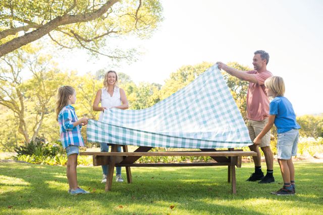 Family spreading tablecloth on picnic table in park, perfect for illustrating outdoor family activities, bonding moments, and leisure time in nature. Ideal for use in advertisements, blogs, and articles about family life, outdoor activities, and summer fun.