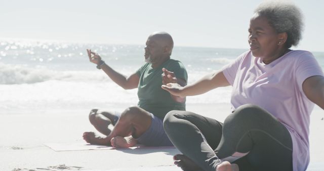 Senior African couple sitting cross-legged on the beach practicing yoga and meditation. Both are focused and appear calm under the bright sunny sky with the ocean waves in the background. This image is ideal for promoting fitness, wellness, and a healthy lifestyle among seniors, as well as portraying the importance of outdoor activities and relaxation routines.