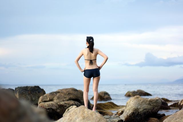 Depicts a young woman in a bikini standing on rocks at the coastline, looking at the ocean with the horizon in the distance. Ideal for concepts related to relaxation, summer vacations, beach activities, or nature exploration. Can be used in travel blogs, wellness magazines, and advertisements for coastal destinations.