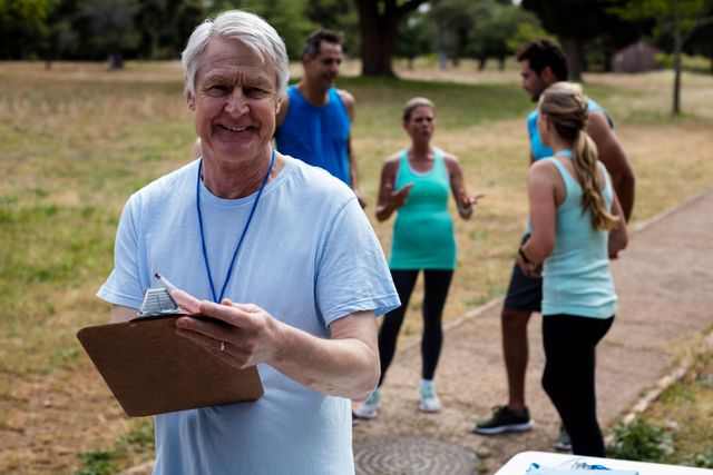Senior male volunteer holding clipboard, smiling in park with group of people in background. Ideal for community service, volunteer work, charity events, team building, and outdoor activities.