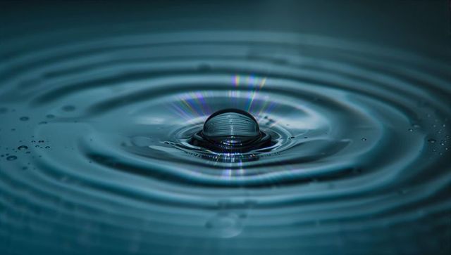 Single water droplet creating concentric ripples with teal reflections and diffraction
