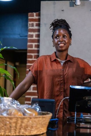 Smiling african woman standing behind counter in cozy brick bakery wearing rust shirt