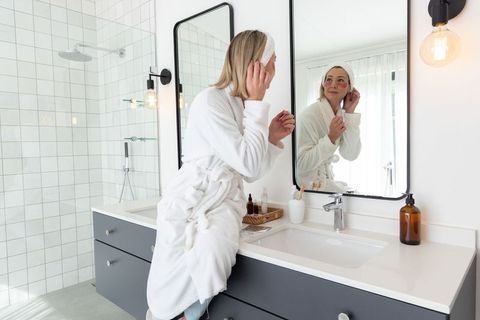 Middle-aged woman applying under-eye patches while sitting on vanity in minimalist bathroom
