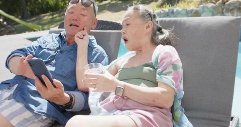 Asian senior couple relaxing poolside, scrolling smartphone and sipping drink on lounger