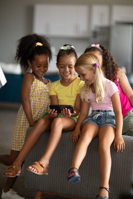 Group of diverse elementary schoolgirls sitting together in a playroom, smiling and using a smartphone. Ideal for concepts related to childhood, education, technology in learning, and friendship. Perfect for educational materials, advertisements for children's products, or articles on the impact of technology on young learners.