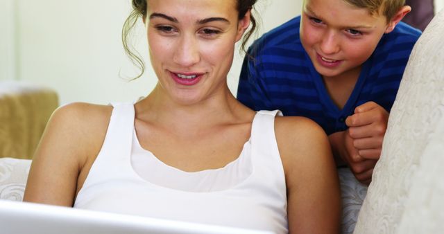 Mother and son engaging with laptop at home. Woman wearing white tank top, boy in blue striped shirt. Ideal for themes related to family, technology use, learning, home life, and bonding moments.