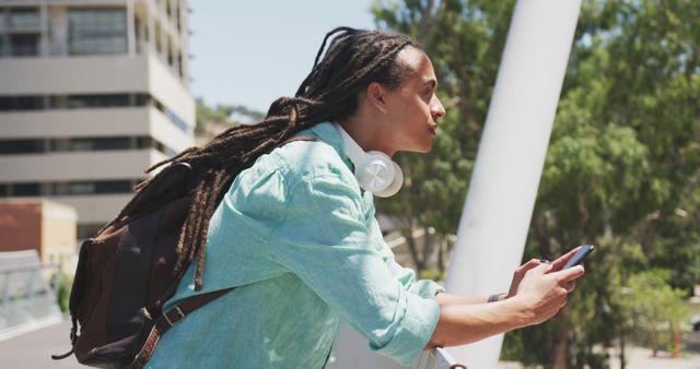 Young man with dreadlocks leaning on railing while holding smartphone. He is also carrying a backpack and headphones around neck. This image can be used for urban lifestyle, technology usage, travel, and youth culture themes.