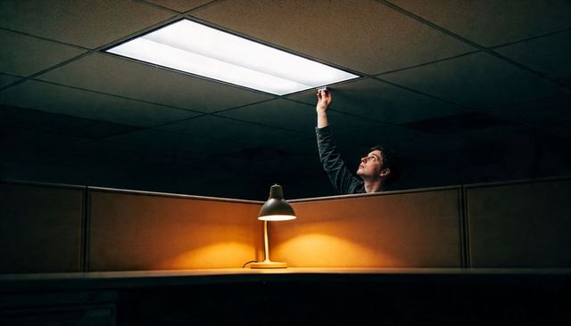 Maintenance worker reaching for fluorescent ceiling panel in dim cubicle with warm desk lamp