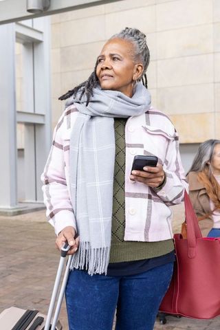 Senior woman tapping phone while pulling suitcase, carrying tote, urban travel, scarf, earrings