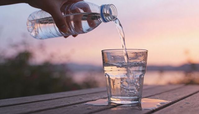 Pouring water from bottle into glass on wooden table at sunset with pastel sky and spill