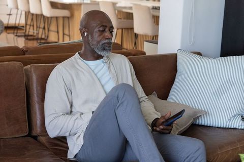 Senior african man relaxing on leather sofa with smartphone and earbud in modern home