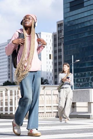 African american woman walking urban crosswalk with headphones and backpack, casual style